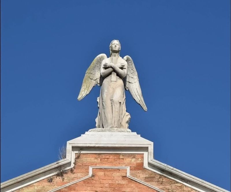 Tradizione del Ponte votivo San Michele Venezia 3 Statua di un angelo con ali in marmo bianco posizionata sopra un edificio in mattoni rossi, cielo azzurro sullo sfondo, vista dall'alto, simbolo di speranza e protezione, elementi architettonici classici, decorazione religiosa.
