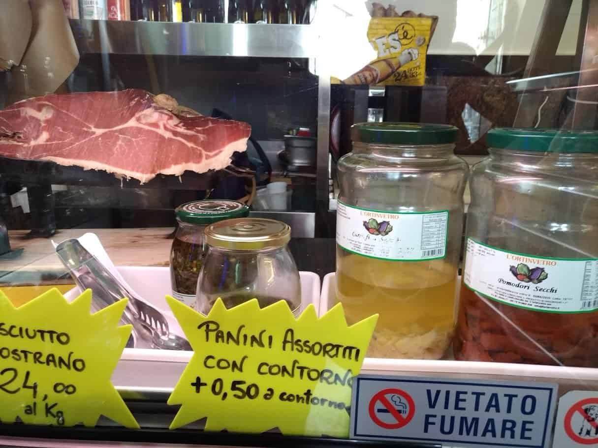 Insaccato stagionato in vetrina con prosciutto crudo e conserve di pomodori secchi e sottaceti, locale alimentare italiano con prezzi e avvisi di divieto di fumo.