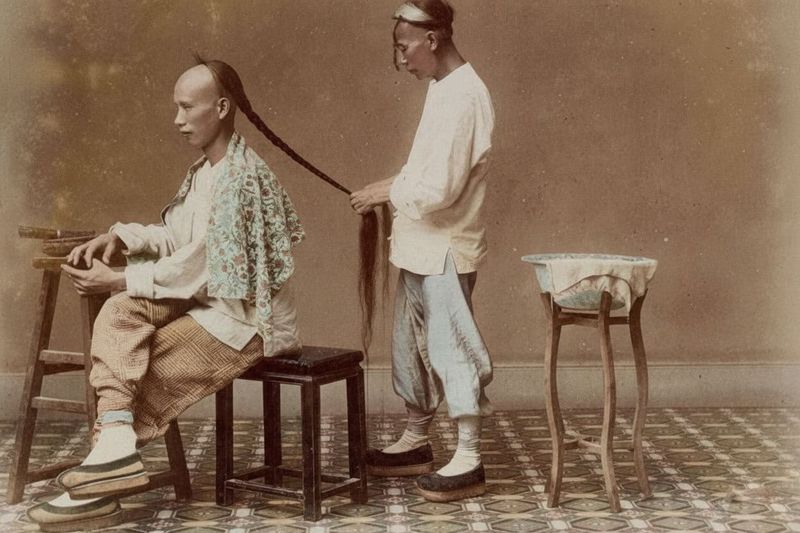 Perché gli uomini della dinastia Qing portavano la treccia manciù? 5 due uomini in abiti tradizionali Qing, uno seduto mentre l'altro intreccia una lunga treccia manciù, interno, stile foto antica, pavimento decorato, atmosfera serena