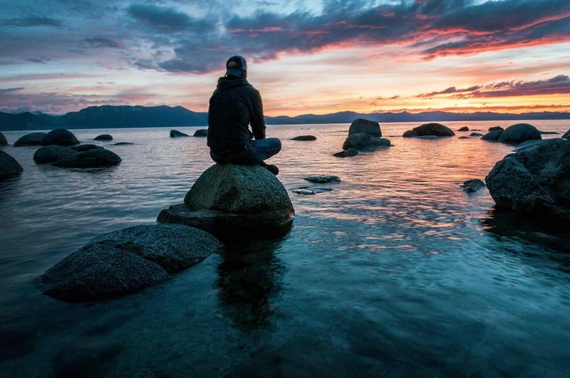 Prenota ora le tue vacanze per trovare le occasioni migliori 3 Riflesso di un tramonto sul lago con persona seduta su una roccia, paesaggio naturale, silenzioso e sereno, ideale per articoli su natura, relax e mindfulness.