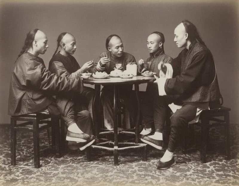 Perché gli uomini della dinastia Qing portavano la treccia manciù? 3 cinque uomini vestiti in abiti tradizionali cinesi Qing, con treccia manciù, seduti attorno a un tavolo rotondo, mentre mangiano e parlano, foto stile antico, interno, tappeto decorato