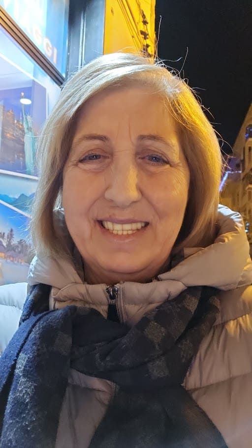 Sorriso di donna anziana con capelli grigi, vestita con cappotto beige e sciarpa scura, in un ambiente urbano notturno con luci colorate e vetrine illuminate.