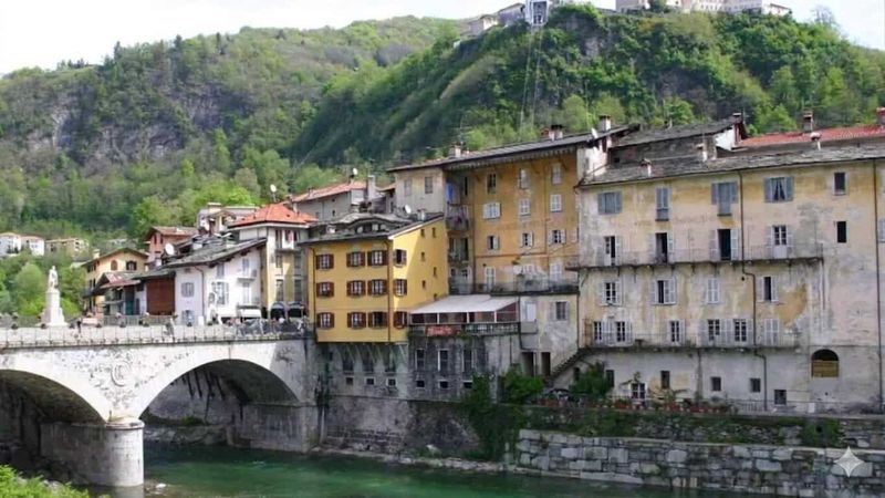 8 attrazioni a Varallo e dintorni in Valsesia 2 Vista di un quartiere antico con edifici colorati e un ponte in pietra che attraversa un fiume, circondato da colline verdi e natura rigogliosa, rappresentando un affascinante esempio di architettura.