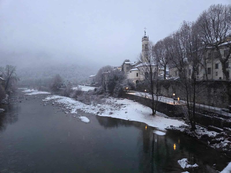8 attrazioni a Varallo e dintorni in Valsesia 12 Paesaggio invernale con il fiume e il centro storico di Varallo Sesia, caratterizzato da edifici storici, una chiesa con torre e alberi ricoperti di neve, ideale per scoprire le attrazioni della zona.