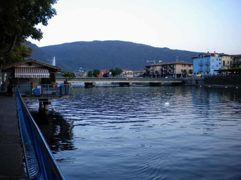 Sarnico cosa vedere: Ville Liberty, lungolago e Cantieri Riva 9 Ponte sul fiume Oglio a Sarnico con vista sulla città e montagne circostanti, ideale per scattare foto panoramiche e godersi il paesaggio naturale e urbano.