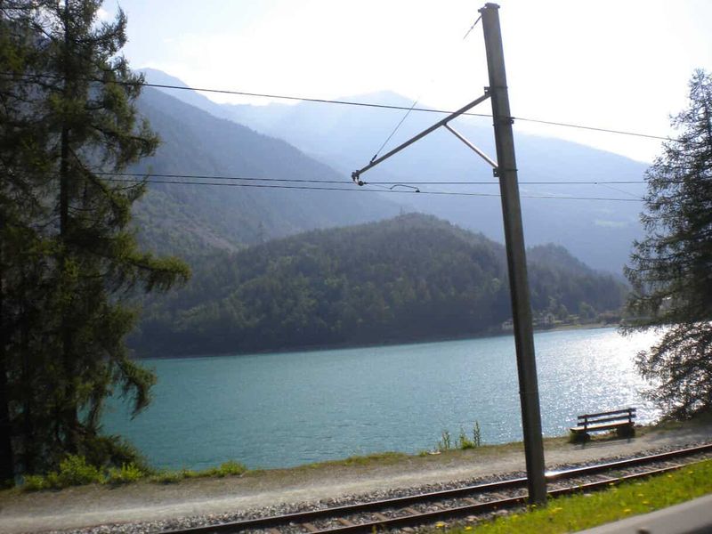Poschiavo Bernina Express, valle italofona Grigioni Svizzera 6 Treno che attraversa un paesaggio naturale con lago e montagne, ideale per appassionati di viaggi e natura. Vista tranquilla e suggestiva di un ambiente alpino con acqua cristallina e vegetazione rigo.