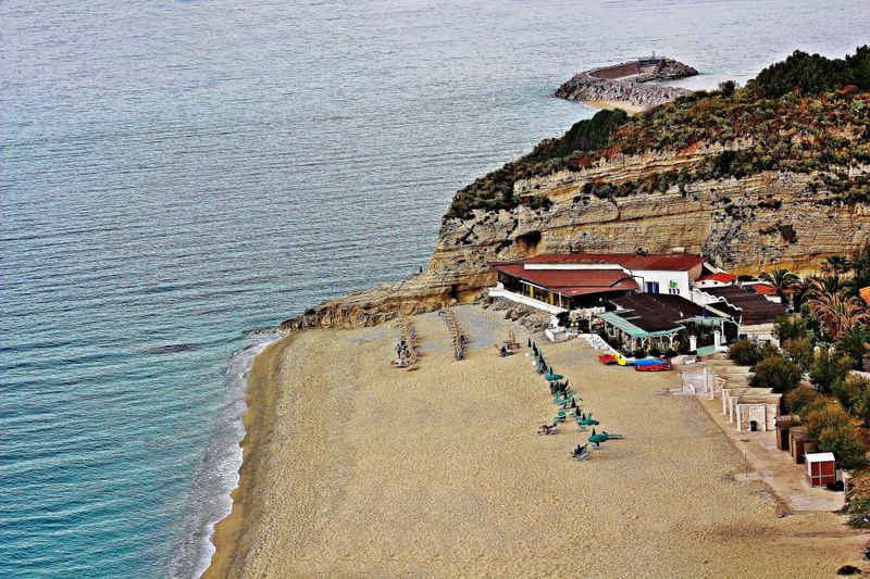 Dalle spiagge dorate della Puglia alle scogliere della Calabria, alla scoperta di un Sud che incanta 2 Spiaggia con sabbia dorata e scogliere, vista sul mare cristallino tra Puglia e Calabria, con stabilimenti balneari e vegetazione mediterranea.