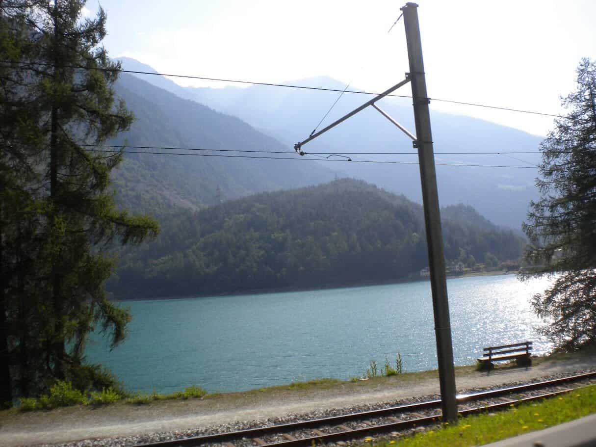 Treno che attraversa un paesaggio naturale con lago e montagne, ideale per appassionati di viaggi e natura. Vista tranquilla e suggestiva di un ambiente alpino con acqua cristallina e vegetazione rigo.
