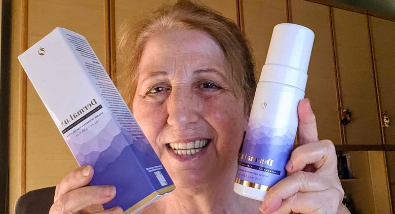 DermaLux Mousse Detergente Viso per pelle matura e spenta 6 Donna sorridente che mostra prodotti per la cura della pelle, crema e lozione, in un ambiente domestico.