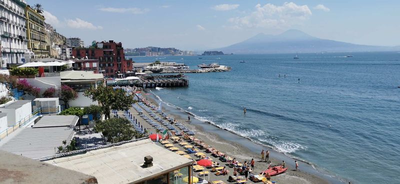Consigli per le vacanze al mare 3 Spiaggia di Napoli con ombrelloni colorati, mare calmo e vista sul Vesuvio sullo sfondo, atmosfera estiva e turistica.
