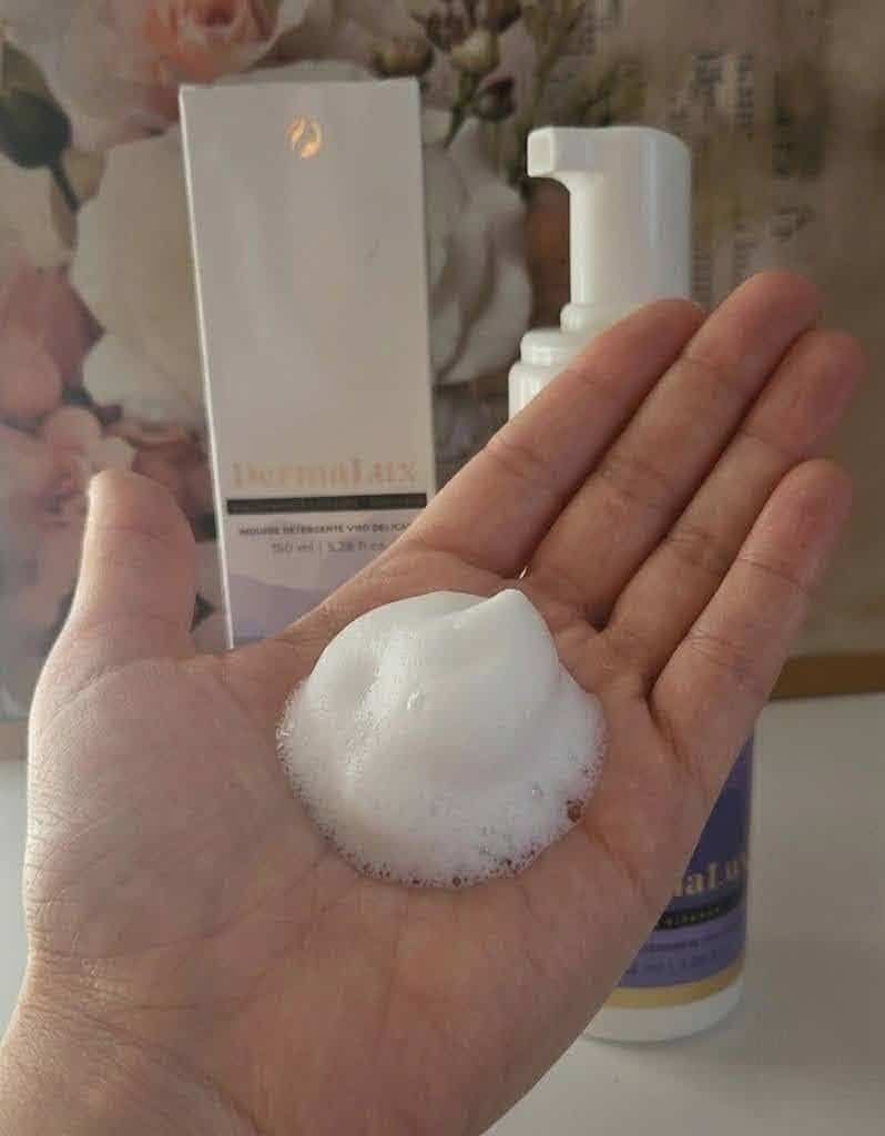 DermaLux Mousse Detergente Viso per pelle matura e spenta 3 Mano con schiuma di un prodotto cosmetico, con flacone e confezione sullo sfondo, focus sulla cura della pelle e prodotti skincare.