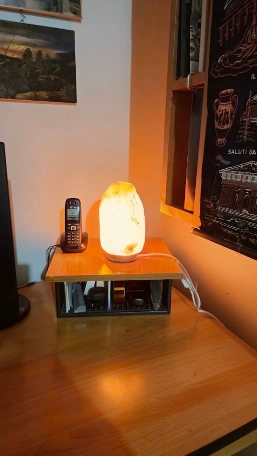 Lampada di sale himalaya: Lampada di sale accesa su tavolo in ambiente domestico, con telefono e decorazioni sullo sfondo, ideale per purificare l'aria in casa.