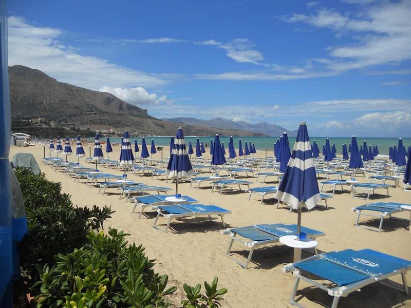 Consigli per le vacanze al mare 4 Sicilia. Spiaggia con numerosi ombrelloni blu e lettini sulla sabbia, vista sul mare e montagne sullo sfondo, cielo sereno con alcune nuvole.