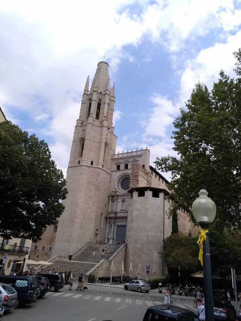 Cosa vedere a Girona in un giorno 3 Impressionante vista della chiesa di Sant'Antonio a Maiorca con la sua torre alta e dettagli architettonici storici, circondata da alberi e un cielo nuvoloso.