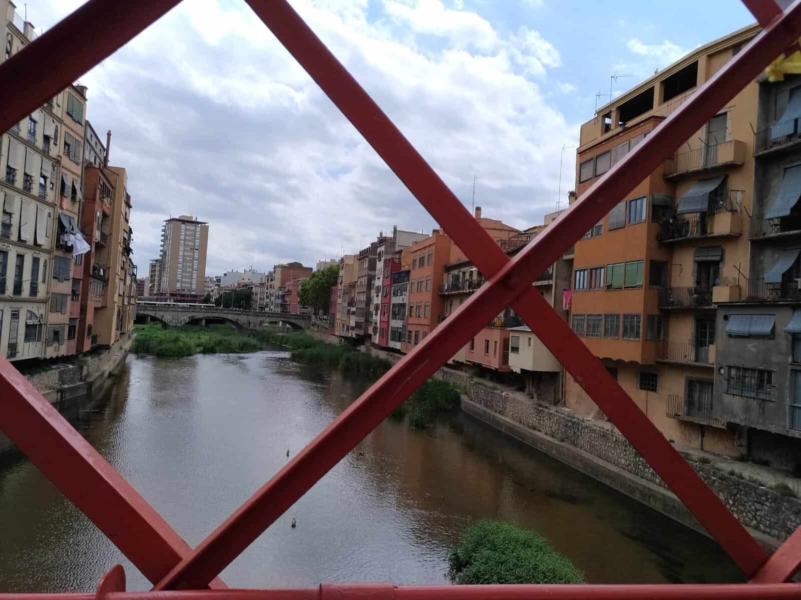 Immagine di un fiume cittadino con edifici residenziali e un ponte rosso in primo piano, cielo nuvoloso, in un contesto urbano.