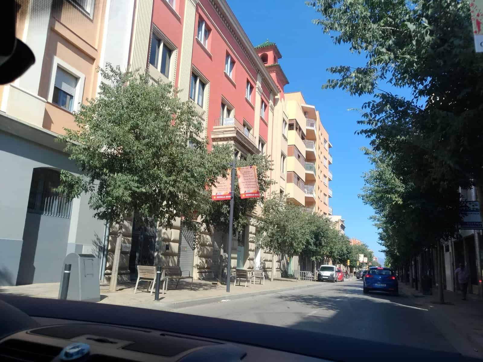 Vista di un vicolo cittadino con edifici colorati e alberi lungo la strada, con auto parcheggiate e un cielo sereno.
