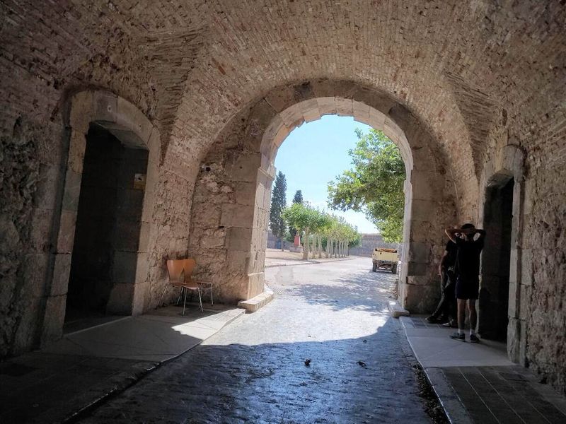 Castello di Sant Ferran a Figueres: la fortezza più grande d'Europa (Orari, Prezzi e Sotterranei) 9 Vista attraverso un arco storico in pietra, con persone che scattano foto e un paesaggio con alberi e strada sotto il cielo azzurro.