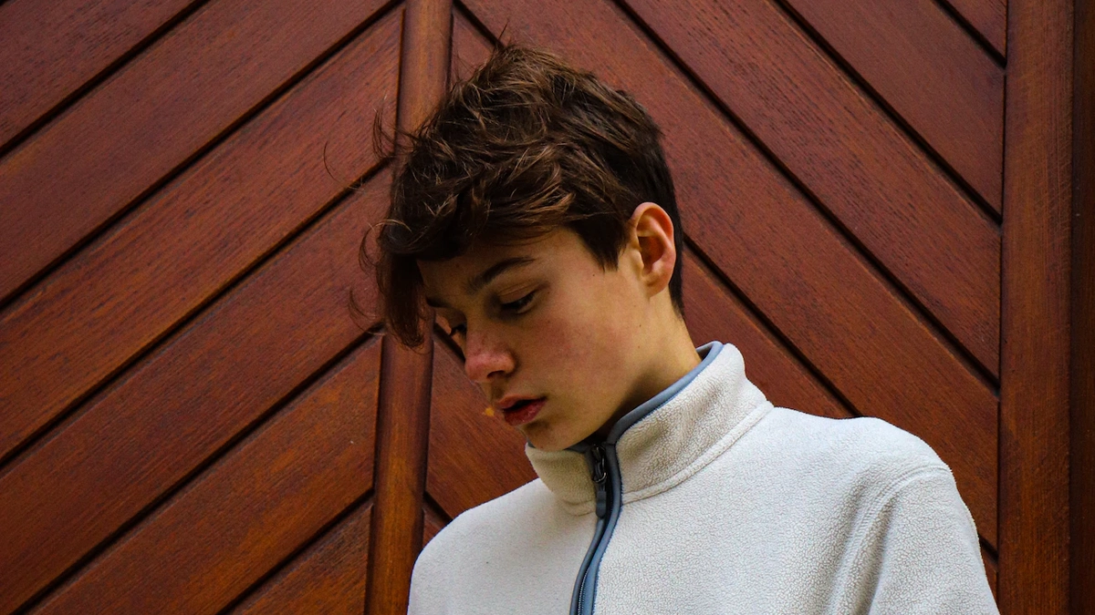 Ergen yaş grubundaki bir erkek, ahşap bir duvar önünde mutsuzca duruyor