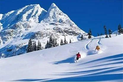 two skiers coming down fresh powder with snow capped mountain in background