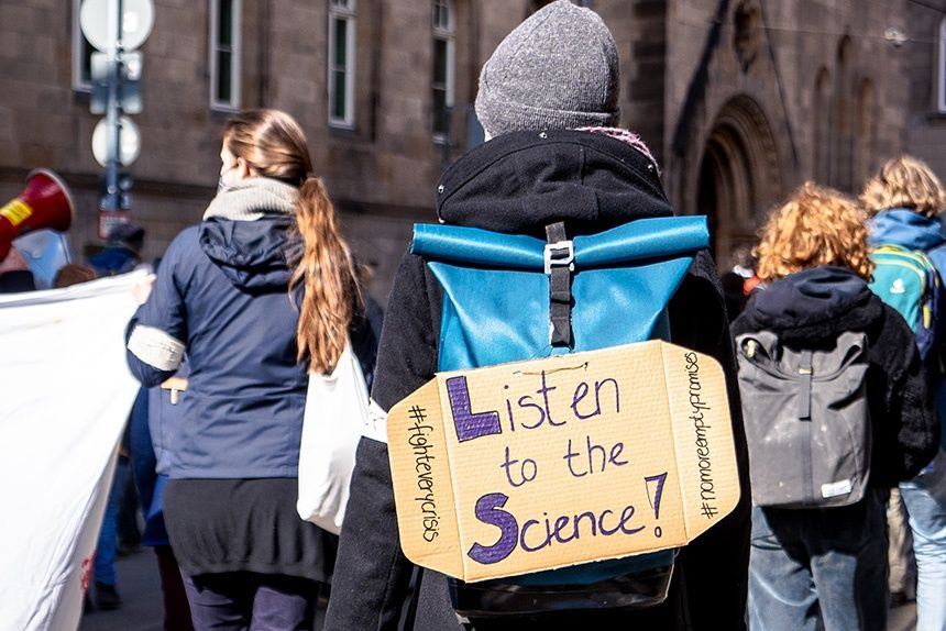 Person am Demo mit Schild am Rücken. Darauf steht 'Listen to the Science!'